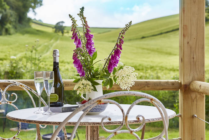 A bottle of prosecco and glasses sit on the outside table at Brownscombe cabin with front row seats of rolling Devon countryside