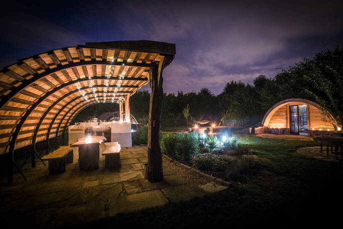 The Secret Garden at Guilden Gate in Hertfordshire under a starry night sky with the hobbit hut and outdoor kitchen illuminated 